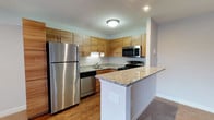 Located at 2617 Dekalb Pike in Norristown, PA, this single-floor space offers a clean, modern aesthetic with thoughtful design throughout. The kitchen serves as a standout feature, showcasing warm wood-toned cabinetry that extends to the ceiling for ample storage, complemented by granite countertops with subtle earth-tone veining. A peninsula counter provides additional workspace and a natural division between the kitchen and adjacent living area. Stainless steel appliances, including a full-size refrigerator, dishwasher, microwave, and range, bring a sleek, contemporary finish to the space. The flooring transitions seamlessly between hardwood-style surfaces in the kitchen and softer carpeting in the living area, offering both practicality and comfort. Recessed lighting and a flush-mount ceiling fixture keep the kitchen bright and well-lit. The overall design emphasizes functionality and modern style, with neutral wall tones creating a versatile backdrop that complements a variety of interior preferences. The open layout between the kitchen and living space enhances the sense of flow and everyday livability throughout this well-maintained single-level residence.