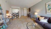 This single-floor residence offers a practical and cohesive layout connecting its principal living spaces. The living room serves as the central gathering area, finished with wall-to-wall carpeting, a soft blue accent wall, and sliding glass doors dressed with sheer white curtains that allow natural light to fill the space. The open sightlines into the dining and kitchen area create a seamless flow throughout the common areas.

The kitchen is equipped with stainless steel appliances, including a gas range, dishwasher, and refrigerator. White cabinetry with dark countertops provides ample storage and prep space, while stone-look tile flooring adds durability underfoot. A breakfast bar with seating connects the kitchen to the adjacent dining area, making it well-suited for both casual meals and entertaining.

The bedroom features carpeted flooring, a window with decorative curtains, and a warm accent wall with vertical paneling detail, contributing a distinct character to the room. A dedicated vanity area with dark countertops is positioned near the bathroom, which includes a shower-tub combination and is complemented by built-in open shelving for convenient storage. Louvered closet doors provide additional organization within the space.