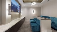 This single-level screening room is thoughtfully designed to deliver a refined, theater-like experience within an intimate setting. The layout features a tiered floor plan, with seating arranged across two levels to ensure optimal sightlines toward the large wall-mounted display screen at the front of the room. Plush teal velvet sofas and armchairs are arranged throughout both tiers, offering comfortable seating while providing a bold pop of color against the room's neutral palette of warm grays and taupe.

The front wall is anchored by a generous flat-screen display, flanked by sleek wall sconces with a linear, textured design that provides ambient lighting along the sides of the room. Beneath the screen, a long run of dark-stained cabinetry is topped with a striking white marble surface featuring elegant veining, adding a luxurious touch. The backsplash behind the counter is clad in a fine metallic mosaic tile, enhancing the room's upscale aesthetic.

Overhead, a tray ceiling with a warm metallic finish houses a central pendant light fixture alongside recessed can lighting. Textured wallcoverings, plush carpeting, and carefully layered lighting throughout contribute to an atmosphere that is both functional and sophisticated.