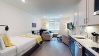 This thoughtfully designed studio unit offers a seamless open-plan layout that efficiently combines living, sleeping, dining, and cooking areas into one cohesive space. Wide-plank hardwood flooring flows throughout, lending warmth and continuity to the interior.

The sleeping area features a queen-sized bed dressed in crisp white linens with yellow accent bedding, flanked by wall-mounted artwork that adds a contemporary touch. Adjacent to it, the living area is anchored by a generous sectional sofa with blue accent pillows, a rustic wood coffee table, and a large wall-mounted flat-screen TV. A compact dining nook with a bistro-style table and chairs provides a functional workspace or casual dining spot.

The kitchen is a standout feature, showcasing a striking two-tone design with navy lower cabinetry and white upper cabinets, topped with clean white countertops and a classic subway tile backsplash. Stainless steel appliances, including a dishwasher and microwave, complete the modern setup.

Natural light filters in through a window overlooking greenery, brightening the crisp white walls and recessed lighting throughout. The bathroom features classic hexagonal mosaic floor tiles, a single vanity with white cabinetry, and a full shower-tub combination, all finished in a clean, neutral palette.