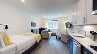 This thoughtfully designed studio unit offers a seamless open-plan layout that efficiently combines living, sleeping, dining, and cooking areas into one cohesive space. Wide-plank hardwood flooring flows throughout, lending warmth and continuity to the interior.

The sleeping area features a queen-sized bed dressed in crisp white linens with yellow accent bedding, flanked by wall-mounted artwork that adds a contemporary touch. Adjacent to it, the living area is anchored by a generous sectional sofa with blue accent pillows, a rustic wood coffee table, and a large wall-mounted flat-screen TV. A compact dining nook with a bistro-style table and chairs provides a functional workspace or casual dining spot.

The kitchen is a standout feature, showcasing a striking two-tone design with navy lower cabinetry and white upper cabinets, topped with clean white countertops and a classic subway tile backsplash. Stainless steel appliances, including a dishwasher and microwave, complete the modern setup.

Natural light filters in through a window overlooking greenery, brightening the crisp white walls and recessed lighting throughout. The bathroom features classic hexagonal mosaic floor tiles, a single vanity with white cabinetry, and a full shower-tub combination, all finished in a clean, neutral palette.