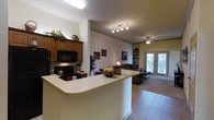 This single-floor 2-bedroom, 2-bathroom residence offers a thoughtfully designed open-concept layout that seamlessly connects the kitchen and living areas. The kitchen is well-appointed with rich wood cabinetry, a black appliance suite including a built-in microwave and gas range, and a wraparound breakfast bar that provides ample prep space and casual dining. Track lighting overhead ensures excellent task illumination throughout the cooking area.

The open floor plan transitions naturally into the living room, where a ceiling fan with light fixture keeps the space comfortable. Built-in shelving provides practical display and storage options, while framed artwork adds a warm, personalized touch to the neutral walls. Light tile flooring runs throughout the main living areas, contributing to a clean and cohesive aesthetic.

Natural light pours in through a set of elegant French doors at the rear of the living room, offering views of the outdoors and creating an inviting, airy ambiance. The overall design balances functionality with comfort, making this residence well-suited for everyday living. The neutral color palette and warm wood accents throughout lend the space a welcoming character that is both practical and aesthetically appealing.