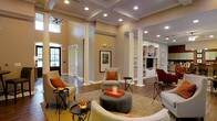 This single-story space makes a striking impression with its soaring ceilings and coffered detailing overhead, lending an air of grandeur to the open-concept layout. Tall decorative white columns frame the main living area, defining zones while maintaining a seamless visual flow throughout the space. Recessed lighting is strategically placed throughout the ceiling, complementing the natural light that filters in through large windows and a glass-paneled entry door near the foyer.

The living area is generously proportioned, anchored by a plush area rug and furnished with a curved sectional sofa, rounded upholstered ottoman, and accent chairs dressed in warm orange throw pillows—adding pops of color against the soft beige walls. Rich hardwood flooring extends throughout, adding warmth and continuity.

A built-in shelving and entertainment unit lines one wall, offering ample display and storage space alongside a media center with arched architectural detailing. Arched passageways echo this design motif, contributing to the cohesive, classical aesthetic. The overall ambiance is refined yet inviting, blending architectural character with comfortable, well-appointed furnishings.