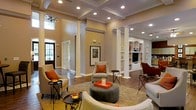 This single-story space makes a striking impression with its soaring ceilings and coffered detailing overhead, lending an air of grandeur to the open-concept layout. Tall decorative white columns frame the main living area, defining zones while maintaining a seamless visual flow throughout the space. Recessed lighting is strategically placed throughout the ceiling, complementing the natural light that filters in through large windows and a glass-paneled entry door near the foyer.

The living area is generously proportioned, anchored by a plush area rug and furnished with a curved sectional sofa, rounded upholstered ottoman, and accent chairs dressed in warm orange throw pillows—adding pops of color against the soft beige walls. Rich hardwood flooring extends throughout, adding warmth and continuity.

A built-in shelving and entertainment unit lines one wall, offering ample display and storage space alongside a media center with arched architectural detailing. Arched passageways echo this design motif, contributing to the cohesive, classical aesthetic. The overall ambiance is refined yet inviting, blending architectural character with comfortable, well-appointed furnishings.