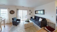 This single-floor residence offers a thoughtfully arranged layout with distinct living, sleeping, dining, and kitchen areas. The living room serves as a welcoming central space, featuring light hardwood flooring, a large sliding glass door that floods the room with natural light and provides direct access to an outdoor deck. A wall-mounted mirror, framed decorative art, and a plush area rug contribute to the room's comfortable, casual ambiance.

The kitchen is well-appointed with warm wood cabinetry, stainless steel appliances including a gas range and refrigerator, and a peninsula with a curved countertop that provides additional workspace and casual seating. Pendant lighting above the peninsula adds a functional yet decorative touch. Adjacent to the kitchen, a dining area offers space for everyday meals.

The bedroom is generously sized, finished with plush carpet, and features a wall-mounted television, a rustic wood-panel media console, and decorative wall art above the bed. A window allows natural light and outdoor views into the space. The bathroom includes tile flooring, a shower with a curtain enclosure, and a vanity with wood cabinetry, a single sink, and well-lit mirror. Overall, the home maintains a neutral, cozy aesthetic throughout.