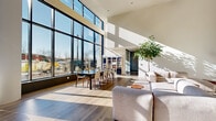 Located at 215 Grove St in Jersey City, NJ, this contemporary communal living space offers a striking single-floor layout defined by dramatic double-height ceilings and expansive floor-to-ceiling glass curtain walls that flood the interior with abundant natural light. The open-plan living room is generously sized, providing distinct seating and gathering zones that flow seamlessly into one another. Large, light-toned hardwood floors span the space, contributing to its bright and airy atmosphere. A comfortable lounge area features plush, neutral-toned sectional sofas and accent chairs arranged around a round wood coffee table, while a separate dining zone accommodates a sizable dark wood table with modern chairs — ideal for group gatherings. An additional casual seating cluster near the glass facade offers an intimate setting with direct views of the surrounding Jersey City streetscape and skyline. Lush indoor trees add organic warmth and a touch of nature to the sleek, modern interior. The architectural design emphasizes clean lines, white walls, and minimalist finishes, creating a polished yet welcoming ambiance. A dedicated conference or meeting table with gold accent lamps sits adjacent to the panoramic windows, completing this versatile and well-appointed communal space.