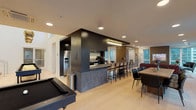 This thoughtfully designed amenity space delivers a seamless blend of functionality and contemporary style across a single, open floor plan. At its core is a fully equipped kitchen anchored by a striking dark-finished island with an integrated cooktop and range, complemented by a separate prep wall featuring stainless steel appliances — including a built-in oven, dishwasher, and refrigerator — sleek dark cabinetry, under-cabinet lighting, and a subway tile backsplash. A generous bar-height counter with seating provides a casual gathering spot adjacent to the kitchen.

The open layout flows naturally into a formal dining area outfitted with a solid wood table and tufted black leather chairs, bathed in natural light from large windows framing urban skyline views. The lounge area offers a striking contrast in textures, with a deep burgundy tufted Chesterfield sofa, a black marble coffee table, and a built-in media wall featuring warm wood paneling, a wall-mounted TV, and a cozy upholstered window nook. Additional seating and a dedicated game area — complete with a pool table and shuffleboard — round out the entertainment offerings. Light wood-look tile flooring runs throughout, tying the spaces together with warmth and cohesion.