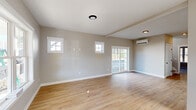 This newly constructed two-floor home showcases clean, contemporary design with a neutral color palette and quality finishes throughout. The main living area is open and airy, featuring warm hardwood-style flooring, exposed ceiling beams, and a wall of large sliding glass doors that flood the space with natural light and provide direct access to the outdoors. Flanking the sliding doors are charming square windows with classic white trim that add architectural character. A wall-mounted mini-split system provides efficient climate control. The kitchen is thoughtfully designed with white shaker-style cabinetry, white subway tile backsplash, light quartz countertops, a central island, and stainless steel appliances including a refrigerator and dishwasher. Stylish pendant lighting anchors the kitchen space. The bathrooms are finished with light tile flooring and crisp white fixtures; one features a combined tub and shower, while another offers a walk-in shower stall, both with natural window light. The bedrooms are well-proportioned, with hardwood-style floors and windows offering exterior views. Throughout the home, white panel doors with matte black hardware add a refined, modern touch, contributing to a cohesive and polished overall aesthetic.
