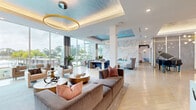 Located at 375 54th Ave N in Saint Petersburg, FL, this sophisticated single-floor amenity space spans a generous footprint and delivers a polished, resort-caliber experience. The grand lobby serves as the centerpiece, boasting soaring double-height ceilings, floor-to-ceiling glass walls, and an abundance of natural light. A sleek reception desk clad in veined stone is flanked by gold-accented open shelving, navy blue accent chairs, and woven pendant light fixtures, creating an inviting yet professional first impression. Throughout the space, light-toned hardwood flooring in a herringbone pattern complements a neutral, coastal-inspired palette accented with deep blues, warm golds, and crisp whites. Multiple lounge areas offer varied seating arrangements, including a sitting area anchored by a dramatic full-height marble feature wall with an integrated linear fireplace and wall-mounted display screen. A grand piano adds a cultural focal point to the open social lounge. A dedicated conference room features large windows overlooking tropical landscaping, a sleek meeting table, and a wall-mounted screen alongside a decorative moss installation. A café-style bar area with dark cabinetry, a white countertop island, gold pendant lighting, and patterned wallcovering provides a functional refreshment hub. Panoramic views of the pool and waterway are visible throughout, enhancing the bright, open, and luxurious ambiance of this exceptional amenity space.