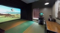 This single-floor indoor golf simulator space is thoughtfully designed to deliver an immersive golf experience regardless of weather or season. The centerpiece of the space is a large-format High Definition Golf projection screen, flanked by floor-to-ceiling blackout curtains that eliminate ambient light and enhance the visual quality of the simulated course imagery. The striking visuals displayed — including coastal links-style courses — create a convincing sense of being on a real fairway.

The hitting area features multi-toned synthetic turf in green, orange, and tan zones, clearly delineating the tee and approach positions. A dedicated control station with a monitor and media shelf sits adjacent to the simulator screen, providing easy access to course selection and performance data. A window with slatted blinds provides natural light control on one side of the simulator bay.

The lounge area is finished with dark grey walls, green carpet, and pendant lighting. A rustic wooden table with dark leather stools serves as a gathering spot, complemented by a bold equestrian canvas print above. Golf bags and additional equipment are neatly stored nearby, completing the functional yet stylish atmosphere of this private golf retreat.