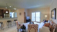 This single-floor residential space offers a comfortable and well-appointed layout designed for everyday living. The living area serves as the heart of the home, featuring plush carpeting, warm neutral-toned walls, and an abundance of natural light streaming in through a set of elegant French doors that open to an outdoor balcony, providing a bright and airy atmosphere.

The living room is thoughtfully arranged with stylish patterned accent chairs and a cozy sofa, centered around a low-profile coffee table. A dedicated media area accommodates a flat-screen television, completing the functional yet inviting setup. Decorative touches, including a sunburst wall clock and a tall bamboo accent piece, add character to the space.

Adjacent to the living area, a pass-through counter connects seamlessly to the kitchen, enhancing the open-concept feel and making the space ideal for entertaining. Pendant lighting above the counter adds a refined touch, while bar-height seating provides a casual dining option.

Overall, the space strikes a balance between practicality and style, with consistent neutral tones, clean lines, and warm lighting creating a cohesive and welcoming ambiance throughout.