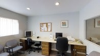 This single-floor space is a compact yet well-appointed computer or business center designed to support productivity and day-to-day office tasks. The room features two dedicated workstations set along an L-shaped or wrap-around light-colored laminate desk with dark wood cabinetry below, providing ample storage through built-in drawers. Each station is equipped with a desktop computer monitor, keyboard, and office chair, allowing multiple users to work simultaneously. A printer and additional peripheral equipment are accessible along the desk surface, making the space fully functional for printing and document management. A cork bulletin board mounted on the wall serves as an informational hub, displaying notices, documents, and updates for occupants or visitors. A framed piece of artwork adds a modest decorative touch to the otherwise utilitarian environment. Recessed ceiling lights provide consistent, even illumination throughout the room. The walls are finished in a neutral light gray tone, contributing to a calm and focused atmosphere. The space is designed with practicality in mind, offering a clean, organized environment suitable for shared use in a residential community, hospitality, or small office setting.