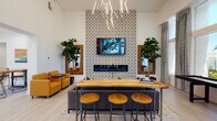 This thoughtfully designed single-floor community lounge space offers a welcoming blend of social and recreational amenities wrapped in a modern, curated aesthetic. The main lounge area immediately draws attention with its dramatic floor-to-ceiling patterned accent wall, which frames a wall-mounted television and sleek linear fireplace below. A sculptural branching chandelier overhead adds an artistic focal point, while warm amber lounge chairs and a live-edge communal bar table with wooden stools create inviting gathering spots throughout.

Light-toned wood-look tile flooring flows seamlessly across the space, complementing the crisp white walls and soaring ceiling heights that amplify the sense of openness. Tall, draped windows flood the game area with natural light and offer views of the surrounding greenery, providing a bright and airy backdrop for the full-length shuffleboard table and adjacent high-top bistro seating.

A dedicated wet bar kitchenette features dark cabinetry with black granite countertops, a stainless steel sink, microwave, and coffee station, making the space fully equipped for entertaining. Curated artwork, decorative mirrors with herringbone wood frames, and lush fiddle-leaf fig plants are thoughtfully placed throughout, enhancing the space's warm yet polished atmosphere. A glass-paneled door leads to an adjoining meeting or flex room, completing this versatile and well-appointed community space.