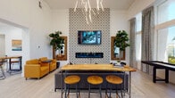 This thoughtfully designed single-floor community lounge space offers a welcoming blend of social and recreational amenities wrapped in a modern, curated aesthetic. The main lounge area immediately draws attention with its dramatic floor-to-ceiling patterned accent wall, which frames a wall-mounted television and sleek linear fireplace below. A sculptural branching chandelier overhead adds an artistic focal point, while warm amber lounge chairs and a live-edge communal bar table with wooden stools create inviting gathering spots throughout.

Light-toned wood-look tile flooring flows seamlessly across the space, complementing the crisp white walls and soaring ceiling heights that amplify the sense of openness. Tall, draped windows flood the game area with natural light and offer views of the surrounding greenery, providing a bright and airy backdrop for the full-length shuffleboard table and adjacent high-top bistro seating.

A dedicated wet bar kitchenette features dark cabinetry with black granite countertops, a stainless steel sink, microwave, and coffee station, making the space fully equipped for entertaining. Curated artwork, decorative mirrors with herringbone wood frames, and lush fiddle-leaf fig plants are thoughtfully placed throughout, enhancing the space's warm yet polished atmosphere. A glass-paneled door leads to an adjoining meeting or flex room, completing this versatile and well-appointed community space.