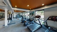 This contemporary fitness center is thoughtfully designed to support a full range of workout routines. The space is defined by a striking two-tone ceiling featuring bold orange and white sections, accented by recessed lighting and ceiling fans that maintain a comfortable atmosphere throughout. Large floor-to-ceiling windows run along the perimeter, flooding the interior with natural light and offering views of the surrounding landscaping.

The main workout floor is generously sized and carpeted in a modern tile-pattern design, housing a comprehensive lineup of cardio equipment including multiple treadmills and elliptical trainers. A dedicated free weights area features a full rack of dumbbells, medicine balls, and exercise benches, complemented by a wall-mounted flat-screen television. A multi-station cable and strength machine anchors the center of the space, catering to a wide variety of strength training needs.

A separate enclosed cycling studio with glass partition walls provides a focused space for spin classes or individual cycling sessions. The facility also includes restroom and changing facilities accessible via a dedicated corridor. Mirrors are strategically placed throughout to enhance the sense of space and support proper form during workouts, making this fitness center a comprehensive and inviting destination for exercise.
