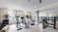 This well-appointed fitness center in Houston's 77007 neighborhood offers a generously sized, single-floor layout designed to accommodate a full range of workout needs. The space is anchored by a robust cardio zone along the windowed perimeter, where treadmills, ellipticals, rowing machines, and stationary bikes are arranged to take full advantage of the large windows overlooking the outdoor pool and palm tree-lined surroundings, providing ample natural light throughout.

Structural columns finished in a soft blue-gray tone punctuate the open floor plan, adding a clean architectural detail that divides the space into functional zones without compromising the open feel. The strength training area features a multi-station cable machine, a free weight rack with dumbbells, and adjustable benches, all positioned alongside floor-to-ceiling mirrors that visually expand the room. A bold, graphic-patterned accent wall adds a striking design element, complementing the neutral tones of the speckled rubber flooring.

Ceiling fans and recessed lighting ensure consistent comfort and visibility throughout the facility. A water fountain station is conveniently mounted on the accent wall. Crown molding along the ceiling ties the aesthetic together, giving the fitness center a polished, well-maintained appearance suited to daily use.