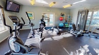 This fitness center offers a comprehensive workout environment designed to support a wide range of exercise routines. The space is finished with soft sage-green walls and durable dark rubber flooring, creating a clean and focused atmosphere conducive to physical activity. Abundant natural light streams in through large windows and glass-paned French doors, which also provide views of the outdoor pool area and surrounding greenery, adding a refreshing ambiance to every workout session.

The facility is well-stocked with cardio equipment, including elliptical trainers, a stair climber, and a stationary bike, all arranged to allow comfortable use and movement throughout the space. A dedicated strength training area features weight machines and benches, catering to resistance and toning exercises. Functional fitness accessories are also on hand, including medicine balls, stability balls, resistance bands, and a rack for organized storage. Framed artwork adorns the walls, adding a touch of warmth to the utilitarian setting.

Overhead fluorescent lighting ensures the space remains bright during all hours. Multiple access points, including interior and exterior doors, provide convenient entry and exit. The center is thoughtfully laid out to maximize usable floor space while maintaining an open, uncluttered feel throughout.
