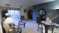 This single-floor residence offers an open and inviting layout that seamlessly combines the living and dining areas into one cohesive space. The living area is anchored by plush carpeting and features a glass-top coffee table, comfortable seating, and a warm floor lamp that contributes to the relaxed ambiance. A striking deep blue accent wall serves as a bold focal point, complemented by dark wood furnishings including a tall armoire and a sideboard topped with a large round mirror, adding depth and sophistication to the interior. The dining area is positioned adjacent to the living space, furnished with a dark wood table and chairs, making it ideal for both casual meals and entertaining. Natural light flows generously into the space through a large sliding glass door, which opens directly onto a private balcony equipped with outdoor seating — offering a pleasant extension of the living area. The neutral toned walls and light carpeting throughout provide a clean, balanced backdrop that allows the decorative elements to stand out. The overall design aesthetic is modern and polished, blending rich, dark wood tones with cool accent colors to create a space that is both functional and visually engaging.