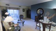 This single-floor residence offers an open and inviting layout that seamlessly combines the living and dining areas into one cohesive space. The living area is anchored by plush carpeting and features a glass-top coffee table, comfortable seating, and a warm floor lamp that contributes to the relaxed ambiance. A striking deep blue accent wall serves as a bold focal point, complemented by dark wood furnishings including a tall armoire and a sideboard topped with a large round mirror, adding depth and sophistication to the interior. The dining area is positioned adjacent to the living space, furnished with a dark wood table and chairs, making it ideal for both casual meals and entertaining. Natural light flows generously into the space through a large sliding glass door, which opens directly onto a private balcony equipped with outdoor seating — offering a pleasant extension of the living area. The neutral toned walls and light carpeting throughout provide a clean, balanced backdrop that allows the decorative elements to stand out. The overall design aesthetic is modern and polished, blending rich, dark wood tones with cool accent colors to create a space that is both functional and visually engaging.