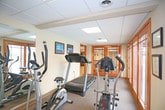 This fitness center is a thoughtfully designed exercise facility offering a comprehensive workout environment within a single-floor layout. The space features a drop-panel ceiling with recessed lighting that ensures consistent, even illumination throughout. A standout feature is the large wall of wood-framed glass doors and windows that floods the room with natural light and provides open views of an adjacent outdoor swimming pool, creating an airy and motivating atmosphere.

The fitness area is equipped with a selection of cardio machines, including a NordicTrack treadmill, an elliptical trainer, and a stationary bike, catering to a variety of exercise preferences. Full-wall mirrors on one side of the room visually expand the space and allow users to monitor their form during workouts. A wall-mounted flat-screen television adds an entertainment option for those exercising. Framed artwork decorates the walls, contributing to a polished and welcoming environment.

Warm wood trim throughout the space complements the neutral tones of the carpeted floors and light-colored walls. A set of elegant wood-framed glass double doors connects the fitness area to an adjacent lounge or gathering space, enhancing the sense of flow and accessibility within the broader facility.