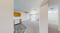 This efficiently designed one-bedroom, one-bathroom unit offers a practical and comfortable single-floor layout. The open-concept living and dining area is finished with plush wall-to-wall carpeting and features a ceiling fan with light fixture, a floor-level HVAC vent, and a sliding glass door flanked by vertical blinds that allows natural light to fill the space. The transition from the living area to the kitchen is marked by a shift to wood-look vinyl plank flooring. The L-shaped kitchen is well-appointed with warm honey-toned oak cabinetry — both upper and lower — white laminate countertops, a double-basin sink, a white electric range with overhead ventilation hood, and a white dishwasher, providing ample storage and functionality. The bedroom is a comfortable size, finished with soft carpeting, white walls, a window with blinds for natural light, and a dedicated closet. The full bathroom includes a single-sink vanity with an oak cabinet base, a large mirror with overhead lighting, wood-look vinyl flooring, and a shower-tub combination with a tiled surround. The unit's clean, neutral palette throughout creates a bright and move-in-ready living environment.