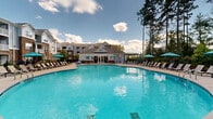 Located at 1090 Summersweet Drive in New Bern, North Carolina, this outdoor amenity space centers around a generously sized, resort-style swimming pool with strikingly clear blue water. The pool's organic, curved shape invites relaxation and leisure, and is complemented by a wide surrounding deck finished in light-toned concrete that provides ample room for sunbathing and socializing.

The deck is furnished with rows of cushioned lounge chairs and teal-accented umbrellas, offering comfortable shaded seating throughout the day. A covered pergola structure provides an additional gathering spot, while a dedicated outdoor grill area supports entertaining. The pool area is fully enclosed by a black wrought-iron fence, with clearly posted pool rules and secure, gated entry points ensuring safety and privacy.

The backdrop of the space features a multi-story residential building constructed with a combination of brick and lap siding, whose balconied units overlook the pool area. On the opposite side, a charming clubhouse with a gabled roof and arched entryway serves as an architectural focal point, framed by tall, mature pine trees that lend a natural, wooded ambiance. Under the open North Carolina sky, this outdoor pool area delivers a welcoming, community-centered retreat ideal for warm-weather enjoyment.