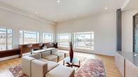 Located along Uptown Ave in Broomfield, Colorado, this single-story communal space showcases a modern, open-plan design with soaring ceilings and an abundance of natural light. Large picture windows line the perimeter, framing sweeping views of the surrounding Broomfield streetscape and open Colorado landscape, flooding the interior with bright, airy light throughout the day.

The main lounge area is anchored by a generous seating arrangement of neutral-toned modular sofas and ottomans, centered around a sleek low coffee table set atop a richly patterned kilim-style area rug. A built-in wood counter with bar-height seating runs along one window wall, offering an ideal workspace or casual perch with an outward-facing view.

Decorative laser-cut metal panels add an artistic focal point near the reception-style counter, which features warm wood cabinetry and a stone-topped surface. Structural columns and a whiteboard panel contribute to the functional character of the space. Light hardwood flooring flows consistently throughout, complementing the clean white walls.

Near the entry, a built-in mailbox bank set against a textured dark accent wall and automatic glass entry doors complete the welcoming, well-appointed lobby experience.