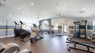 Located at 279 Lindenwood Drive in Exton, PA, this fitness center offers a generously sized, single-floor exercise room designed to support a comprehensive workout experience. The space features soaring vaulted ceilings that lend an open, airy atmosphere, complemented by recessed lighting, ceiling fans, and large windows — including a distinctive arched window — that invite natural light throughout.

The interior is finished with durable rubber flooring in a speckled gray tone, well-suited for high-traffic athletic use. Walls are painted in a two-tone gray scheme accented by a yellow horizontal stripe, giving the space a clean, modern aesthetic. Full-length mirrors along one wall enhance the sense of depth and allow users to monitor their form during workouts.

The cardio zone is equipped with multiple treadmills, elliptical machines, a StairMaster, and stationary bikes, all positioned for ease of use. Wall-mounted flat-screen TVs are available for entertainment during cardio sessions. The strength training area features a multi-station cable machine, weight benches, free weights with a dedicated dumbbell rack, and a variety of resistance equipment. A separate room provides additional utility space. The facility is well-organized, thoughtfully laid out, and suited for a wide range of fitness goals.