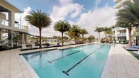 This outdoor amenity space showcases a generously sized lap pool as its centerpiece, finished with crisp turquoise water that reflects the open sky above. The pool is clearly delineated with lap lanes, making it suitable for both recreational swimming and structured exercise. Flanking the pool on either side is a broad, paved sun deck furnished with rows of modern lounge chairs, offering ample space for relaxation and sunbathing. Tall, mature palm trees are positioned throughout the deck, providing a lush tropical ambiance while framing sweeping views of the surrounding landscape. In the background, additional seating areas and pergola-style shade structures are visible, along with what appears to be a waterfront or marina setting, suggesting a prime waterside location. The overall design is clean and contemporary, with a neutral color palette that complements the natural surroundings. The wide-open sky and abundant natural light enhance the bright, airy atmosphere of this well-appointed outdoor retreat, making it a standout feature ideally suited for warm-weather enjoyment.