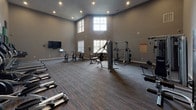 This fitness center in Houston, TX (77049) offers a comprehensive workout environment designed to support a wide range of exercise routines. The space is defined by soaring ceilings and large upper-level windows that flood the interior with natural light, creating an open and energizing atmosphere. The flooring transitions between striped carpet in the main workout area and hard flooring near the cardio zone, providing both comfort and functionality.

A striking feature wall adorned with a bold geometric pattern of black, gray, and green acoustic panels serves as a dynamic backdrop for a row of Spirit treadmills and elliptical trainers, each equipped with individual media screens for entertainment during workouts. Additional cardio options include stationary cycling bikes arranged near a wall-mounted flat-screen television.

The strength training area is well-appointed with a multi-station cable machine, a Smith machine, weight benches, a dumbbell rack, and rowing machines. Stability balls and other functional fitness accessories are neatly stored along the walls, offering variety for flexibility and core training. Double glass-panel doors provide access to an adjacent space, further enhancing the facility's connectivity. Recessed lighting and neutral gray walls complete the clean, modern aesthetic throughout.