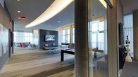 This contemporary amenity lounge is a thoughtfully designed social space that blends sleek architectural details with a welcoming, high-end atmosphere. The standout feature is the dramatic curved ceiling, which is accented by integrated ambient strip lighting that casts a warm, soft glow across the entire room, adding depth and a sense of sophistication to the space.

The open floor plan accommodates a variety of leisure activities, highlighted by a central game area that includes a ping pong table and a billiards table, making it an ideal gathering spot. A large wall-mounted flat-screen television is set against a bold, dark accent wall, creating a natural focal point for relaxed viewing and entertainment.

Substantial concrete cylindrical columns punctuate the space, contributing an industrial character that contrasts elegantly with the polished, modern finishes. The flooring features a striped, neutral-toned carpet that complements the overall color palette of grays, blues, and whites.

Perhaps the most striking element is the abundance of natural light that floods the interior through expansive floor-to-ceiling windows lining the perimeter. These windows frame panoramic views of the sky and surrounding skyline, evoking a sense of elevation and openness. Sheer curtains soften the light while maintaining the airy, luminous quality of the lounge.