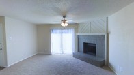 This single-floor space centers around a comfortable living area defined by neutral-toned carpet and a textured popcorn ceiling. A ceiling fan with an integrated light fixture is positioned centrally overhead, providing both airflow and ambient lighting. One of the room's standout features is the full brick fireplace, accented by a white-painted chevron-patterned wood surround that adds a decorative architectural touch. The raised brick hearth extends the fireplace's visual presence into the room. Natural light enters through a large sliding glass door dressed with vertical blinds, offering an outdoor connection while maintaining privacy. The walls are painted in a soft, neutral tone that complements the room's understated palette. The overall ambiance is functional and classic, with a layout that encourages relaxed, everyday living. The combination of the fireplace as a focal point, the ceiling fan for comfort, and the sliding glass door for light and access gives the space a well-rounded, livable character suited for a range of furnishing styles.
