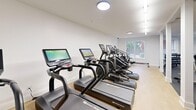 Located at 2025 Renaissance Park Place in Cary, NC, this fitness center offers a thoughtfully organized exercise space across a single floor. The room is divided into two distinct functional zones, separated by a row of white structural columns that lend an architectural rhythm to the interior.

The cardio zone features light wood-style flooring and is outfitted with a lineup of Star Trac treadmills and elliptical machines, each equipped with individual digital display screens. A window at the far end of this area allows natural light to filter in, providing an outdoor view of surrounding greenery to energize workouts.

The strength training zone transitions to dark rubber flooring, providing a durable and supportive surface. This area includes a multi-functional cable machine, a variety of selectorized strength machines, and a full rack of dumbbells. Large mirrors along the walls amplify the sense of space and allow users to monitor their form. A rowing machine is also visible in the layout.

Throughout the space, linear LED lighting and flush-mounted ceiling fixtures provide bright, even illumination. The walls are finished in crisp white, contributing to a clean, modern aesthetic. The overall design is functional and efficient, offering a comprehensive fitness environment well-suited to a range of exercise routines.