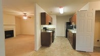 This single-story space is designed with practicality and functionality in mind. The kitchen serves as a central feature, offering a galley-style layout with rich, dark-stained wood cabinetry that provides generous upper and lower storage. Dark laminate countertops run along both sides of the kitchen, offering ample workspace for meal preparation. The kitchen is equipped with a gas range, a refrigerator, and a dishwasher, making it well-suited for everyday cooking needs. A sink with cabinetry on either side is positioned toward the end of the kitchen, completing the practical layout. The flooring in the kitchen consists of neutral-toned tile that is both durable and easy to maintain. The kitchen flows naturally into the adjacent living area, which transitions to carpeted flooring and is illuminated by a ceiling fan with an integrated light fixture, providing both comfort and air circulation. The open connection between the kitchen and living area creates a cohesive, accessible floor plan. The space is finished with white walls and standard baseboards, contributing to a clean and straightforward aesthetic throughout.