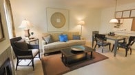This two-storey space offers a well-designed interior with a comfortable and cohesive aesthetic. The living area serves as a central gathering space, furnished with a generously sized beige sofa accented by decorative throw pillows in shades of blue and grey. A dark-finished coffee table anchors the seating area atop a warm brown area rug, adding contrast to the light-toned carpeted flooring beneath. A large framed artwork featuring a circular motif in gold tones provides a striking focal point on the wall above the sofa, contributing to the room's refined yet relaxed atmosphere. Ambient lighting from a classic floor lamp and a side table lamp creates a warm, welcoming glow throughout the space. Adjacent to the main seating area, a dining arrangement with dark wood chairs offers a seamless transition between living and dining functions. The overall palette of warm neutrals, offset by carefully chosen blue accents, lends the space a timeless and balanced character. Spread across two floors, the layout provides a sense of depth and versatility, making it well-suited for both everyday living and entertaining.