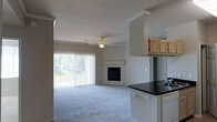 This single-floor space offers an open-concept layout that seamlessly connects the kitchen and living areas, creating a cohesive and functional environment. The living room is finished with plush wall-to-wall carpeting that adds warmth and comfort underfoot, while crown molding along the ceiling lends a polished architectural detail throughout. A built-in fireplace serves as a natural focal point, providing both aesthetic appeal and a cozy atmosphere. A ceiling fan with a light fixture is centrally positioned, ensuring year-round comfort and ambient lighting. Large sliding glass doors dressed with vertical blinds flood the living area with natural light and offer views of the surrounding greenery, with direct access to an outdoor balcony or patio. The kitchen is positioned adjacent to the living space and features dark countertops that contrast neatly with light wood upper cabinetry, providing ample storage. A built-in sink and white appliances complete the kitchen's practical design. The open pass-through between the kitchen and living room enhances the sense of space and allows for easy interaction between the two areas, making this home well-suited for both everyday living and entertaining.