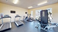 Located at 4020 Eight Belles Ln. in Greensboro, NC, this single-floor fitness center offers a clean, functional environment designed to support a well-rounded workout routine. The room features warm, light-yellow walls and durable rubber flooring, creating a practical and comfortable space for exercise.

Natural light flows in generously through multiple large windows fitted with white plantation shutters, providing views of the surrounding outdoor area. The bright, open feel is further enhanced by overhead fluorescent lighting, ensuring the space remains well-lit throughout the day.

The cardio section is equipped with multiple treadmills, elliptical trainers, and a recumbent bike, each positioned for easy access. Multiple wall-mounted flat-screen televisions are strategically placed throughout the room, allowing users to stay entertained during workouts. A full multi-station cable weight machine provides options for strength training, while a freestanding dumbbell rack offers a range of free weights for varied exercises.

A large floor-to-ceiling mirror spans one wall, visually expanding the space and allowing users to monitor their form. A direct exterior door provides convenient access to the outdoors. The space is monitored by a security camera, and a waste receptacle is provided for user convenience. Overall, this fitness center delivers a comprehensive and inviting environment for daily exercise.