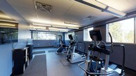 Located at 1600 West Lane Avenue in Upper Arlington, Ohio, this fitness center offers a dedicated space for health and wellness. The room is finished with a drop-panel ceiling fitted with bright fluorescent lighting, ensuring the area is well-lit throughout. Dark rubberized flooring provides a durable, safety-conscious surface suitable for a range of exercise activities. Large windows line the walls, inviting natural light into the space and offering views of the surrounding greenery, creating an open and energizing atmosphere. The facility is equipped with a variety of cardio machines, including ellipticals and a stationary bike, as well as free weight equipment and benches to support both cardiovascular and strength training workouts. The walls are finished in a deep charcoal tone, giving the space a modern and focused aesthetic. Full-length mirrors are positioned along the walls to assist users with proper form and technique. The single-floor layout is thoughtfully organized to allow for comfortable movement between equipment, making efficient use of the available space. Overall, the fitness center provides a functional and well-appointed environment for residents or building occupants to maintain an active lifestyle.