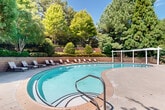 This inviting outdoor amenity space centers around a generously sized freeform swimming pool with crystal-clear turquoise water, designed to evoke a resort-like atmosphere. The pool's curved, organic shape is complemented by elegant tile detailing along its interior, and easy-access stainless steel handrails provide convenient entry. Surrounding the pool is a broad concrete and wood-deck patio, offering ample space for lounging and socializing. A row of cushioned chaise lounge chairs lines one side of the pool, positioned against a tiered retaining wall accented with neatly manicured shrubs and lush greenery. On the opposite side, a white pergola provides a shaded retreat, adding architectural interest to the outdoor setting. Adjacent to the pool, a well-appointed single-story clubhouse features warm yellow exterior walls, a pitched roof, and charming French doors that open directly onto the patio. Multiple patio umbrella tables and chairs are arranged outside the clubhouse, creating a comfortable area for outdoor dining and relaxation. The entire space is enveloped by a mature canopy of tall evergreen and deciduous trees, offering natural shade and privacy while enhancing the serene, park-like ambiance. The combination of thoughtful landscaping, functional amenities, and attractive architecture makes this outdoor pool area an exceptional recreational destination.