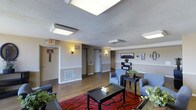 Located at 2038 South Vaughn Way in Aurora, Colorado, this single-floor space presents a well-appointed interior designed for comfort and functionality. The main lounge area serves as a welcoming gathering space, featuring hardwood-style flooring complemented by a bold red patterned area rug that anchors the seating arrangement. A pair of grey upholstered armchairs and a dark-finished coffee table create an inviting conversational setting, accented by potted greenery that adds a touch of warmth. Overhead, flush-mounted fluorescent lighting and a textured ceiling provide consistent, even illumination throughout. Wall-mounted sconces along the corridor hallways offer softer accent lighting, contributing to a balanced and comfortable ambiance. The space includes multiple doorways and corridor access points, suggesting a multi-room or multi-unit layout extending beyond the central lounge. Neutral-toned walls with a contrasting lower wainscoting detail lend a clean, structured aesthetic. A landscape photograph mounted on the wall adds a decorative touch reflective of the Colorado setting. Safety signage, including clearly marked exit indicators, ensures the space meets practical building requirements. Overall, the space combines accessibility, comfort, and a functional design suitable for communal or hospitality use.