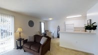 This single-floor residence is designed with an open, connected layout that promotes a sense of spaciousness and ease of movement. The living area is carpeted throughout and serves as the central gathering space, featuring a large sliding glass door dressed with vertical blinds that invites abundant natural light and provides views of the outdoors. The room comfortably accommodates seating and entertainment areas, with neutral wall tones that create a clean, welcoming atmosphere and a textured ceiling that adds subtle depth overhead.

Adjacent to the living room, the dining area flows naturally from the main space, positioned near a window that further brightens the interior. The compact yet functional kitchen is separated by a peninsula-style countertop that opens to the living and dining areas, creating a convenient pass-through design. White cabinetry and matching appliances, including a full-sized refrigerator, contribute to a bright, cohesive aesthetic. The open sightlines between the kitchen, dining, and living zones make this residence feel notably airy and interconnected. Overall, the space presents a practical and comfortable single-level layout with a light, neutral palette throughout.