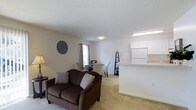 This single-floor residence is designed with an open, connected layout that promotes a sense of spaciousness and ease of movement. The living area is carpeted throughout and serves as the central gathering space, featuring a large sliding glass door dressed with vertical blinds that invites abundant natural light and provides views of the outdoors. The room comfortably accommodates seating and entertainment areas, with neutral wall tones that create a clean, welcoming atmosphere and a textured ceiling that adds subtle depth overhead.

Adjacent to the living room, the dining area flows naturally from the main space, positioned near a window that further brightens the interior. The compact yet functional kitchen is separated by a peninsula-style countertop that opens to the living and dining areas, creating a convenient pass-through design. White cabinetry and matching appliances, including a full-sized refrigerator, contribute to a bright, cohesive aesthetic. The open sightlines between the kitchen, dining, and living zones make this residence feel notably airy and interconnected. Overall, the space presents a practical and comfortable single-level layout with a light, neutral palette throughout.