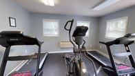 This fitness center is a single-floor facility designed to provide residents with a convenient and functional space for exercise. The interior is finished with cool gray walls and durable rubber flooring, creating a clean, practical environment suited for physical activity. Flush-mounted fluorescent lighting ensures the space is well-lit throughout.

The cardio area is the centerpiece of the facility, housing multiple Sole-brand treadmills and an elliptical trainer, offering a variety of low- and high-impact cardiovascular workout options. A stationary upright bike adds further versatility for cardio training. A multi-station cable strength machine provides users with options for resistance and strength training exercises. A wall-mounted television is positioned to keep users entertained during workouts.

Natural light filters in through several windows fitted with horizontal blinds, offering views of the surrounding greenery. A wall-mounted HVAC unit ensures year-round climate comfort. The facility is accessible through a set of glass-paneled double doors that open to the outdoors. A bulletin board and whiteboard near the entrance provide a space for notices and information. A fire extinguisher is also on-site, reflecting attention to safety standards throughout the facility.