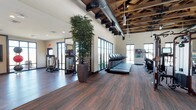 This fitness center combines industrial design with an inviting, open atmosphere. The space is anchored by striking exposed wooden beam ceilings with visible ductwork, lending the room a raw, warehouse-inspired aesthetic that is both functional and visually dynamic. Pendant lighting and ceiling fans complement the abundant natural light that pours in through large, dark-framed floor-to-ceiling windows lining the walls, creating a bright and energizing environment throughout the day.

The flooring features rich, dark wood-toned planks that add warmth and contrast to the lighter walls, tying the industrial and natural design elements together seamlessly. A row of treadmills and cardio machines is positioned along the windowed wall, allowing users to enjoy outdoor views during their workouts. Adjacent to this, a dedicated strength and resistance training zone houses a selection of weight machines and equipment, partially separated by a mirrored partition that enhances the sense of space.

Tall decorative bamboo plants are strategically placed throughout, introducing a touch of nature and tranquility to the active environment. The generous ceiling height and open floor plan allow for comfortable movement and spacing between equipment. Colorful artwork adorns one wall, adding a creative accent to the space. Overall, this fitness center offers a well-rounded, thoughtfully designed setting for a complete workout experience.