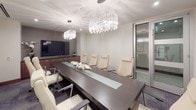 This elegantly appointed conference room delivers a polished, professional environment designed for high-level meetings and presentations. At the center of the space sits a substantial dark wood conference table with a frosted glass inlay runner, surrounded by cream-colored high-back leather executive chairs that comfortably accommodate a sizeable group of attendees. Two striking crystal chandeliers suspended from the smooth white ceiling serve as the room's focal point, casting a warm, sophisticated glow throughout the space. Along one wall, a rich walnut credenza with dark granite countertop houses a large flat-screen display, providing seamless presentation capabilities. The credenza's ample cabinetry offers generous storage while maintaining a clean, uncluttered aesthetic. Curated abstract artwork in monochromatic tones adorns the walls, complementing the room's refined contemporary palette of warm neutrals and deep wood tones. Patterned commercial carpeting in muted blue-grey tones grounds the space with subtle visual interest. A frosted glass panel door provides privacy while maintaining a sense of openness. The overall ambiance is one of understated luxury and functional elegance, making this conference room well-suited for executive gatherings, client presentations, and collaborative business sessions.