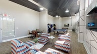 This dynamic recreational space combines entertainment and relaxation in a single open-plan layout designed for socializing and play. The room is defined by its bold industrial aesthetic, featuring a dramatic dark-painted ceiling with exposed track lighting and pendant fixtures that cast a warm, energetic glow across the space. Light wood-style flooring runs throughout, complementing the neutral grey walls and providing a clean, modern foundation.

The lounge area anchors one end of the room, furnished with vibrant chevron-patterned chairs and sofas in a striking mix of red, blue, and white tones. These seating clusters are arranged around low white coffee tables, creating inviting spots to unwind. A bold, graphic mural of intersecting black lines decorates one wall, adding an artistic focal point alongside a wall-mounted flat-screen TV. High windows along this wall allow natural light to filter in and offer views of the surrounding cityscape.

The entertainment zone occupies the opposite side of the room, housing a full-size ping-pong table, a foosball table, and a collection of classic upright arcade machines — including Space Invaders — evoking a retro gaming atmosphere. An additional wall-mounted TV provides further viewing options. A small work or dining nook with a round table and ergonomic chairs rounds out the space, ensuring functionality alongside fun.