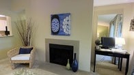 This single-floor space offers a thoughtfully designed interior that balances comfort and style. The living area serves as the heart of the space, centered around a tile-surround fireplace that provides a classic focal point. Above the fireplace, a bold blue abstract artwork adds a vibrant splash of color against the neutral, warm-toned walls, creating a visually striking contrast. Decorative vases — one in deep navy and another in sage green — are neatly arranged on the fireplace hearth, contributing to the curated aesthetic of the room. The flooring is carpeted, lending a soft, comfortable feel underfoot throughout the living area. Natural and ambient lighting work together to maintain a warm and welcoming atmosphere. A glimpse into an adjacent area reveals additional living or workspace, complete with a dark console or desk and a mirror that enhances the sense of depth and openness. Woven basket accents and dried botanical arrangements add organic texture to the décor. The overall design aesthetic leans toward a calm, contemporary style with neutral tones and carefully selected decorative elements that make the space feel both lived-in and polished.