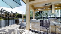 This single-story property showcases a well-appointed covered outdoor entertaining space designed for comfortable al fresco living. The covered patio is supported by substantial stucco-finished columns with arched detailing, lending a refined architectural character to the exterior. A ceiling fan is mounted overhead, providing airflow during warm weather, while recessed and pendant-style lighting ensure usability into the evening hours.

A standout feature of this outdoor area is the built-in bar, constructed with stacked natural stone cladding and topped with a granite countertop. Bar-height seating invites casual dining and entertaining directly at the counter. Additional woven strap patio chairs complete the seating arrangement, offering a relaxed and functional layout.

The flooring consists of large-format stone or tile pavers that extend throughout the covered and open patio areas, contributing to a cohesive, upscale aesthetic. A black wrought iron fence defines the perimeter, providing security while preserving unobstructed views of the surrounding landscape. Mature palm trees, lush greenery, and what appears to be a pond or water feature in the background create a serene, tropical ambiance that enhances the overall outdoor living experience.