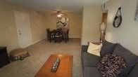 This single-floor residence offers an open and cohesive layout that seamlessly connects the main living and dining areas. The living room is anchored by a plush, gray sofa accented with decorative pillows, complemented by a warm wooden coffee table that adds a touch of natural character to the space. Soft, neutral-toned carpet flows throughout, contributing to the overall warmth and comfort of the interior.

The dining area is positioned directly adjacent to the living room, creating an effortless transition between the two spaces. A dark wood dining table paired with matching chairs serves as the focal point of the dining zone, while a bold floral wall art piece provides a striking decorative accent. A ceiling fan with integrated lighting overhead offers both practical airflow and ambient illumination.

Just beyond the dining area, a glimpse into the kitchen reveals cabinetry and countertop space, suggesting a functional and accessible cooking area. The textured ceiling and neutral wall tones throughout lend the space a classic, understated aesthetic. Overall, this residence presents a practical and inviting single-floor layout well-suited for comfortable everyday living.