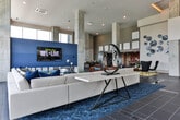 Located at 151 Mills St NW in Atlanta's dynamic Westside, this contemporary amenity lounge delivers an impressive blend of modern design and functional gathering spaces. The space is defined by soaring ceilings and expansive floor-to-ceiling windows that flood the interior with natural light and frame panoramic views of the Atlanta skyline and surrounding greenery. Exposed concrete columns punctuate the open layout, lending an industrial-chic character that contrasts beautifully with the refined furnishings throughout. The main lounge area features a generous sectional sofa and plush burnt-orange accent chairs arranged around a bold blue patterned rug, anchored by a wall-mounted flat-screen TV set against a striking blue textured feature wall. A dedicated billiards area showcases a full-size pool table positioned to take full advantage of the sweeping city views through the glass curtain wall. A large communal high-top table with ample seating provides an ideal space for collaborative gatherings or casual dining, complemented by direct access to an outdoor terrace. Curated artwork and sculptural wall installations add a gallery-like quality throughout, while a warmly lit dining nook with pendant lighting and brick accents offers an intimate contrast to the airy main hall. Large-format tile flooring and polished concrete surfaces complete the sophisticated, cohesive aesthetic of this versatile community space.